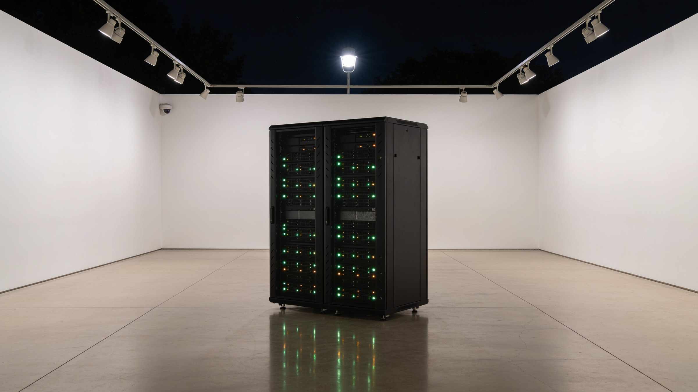 At night in an empty gallery the server rack glows with small green and amber lights, reflected in the polished floor.