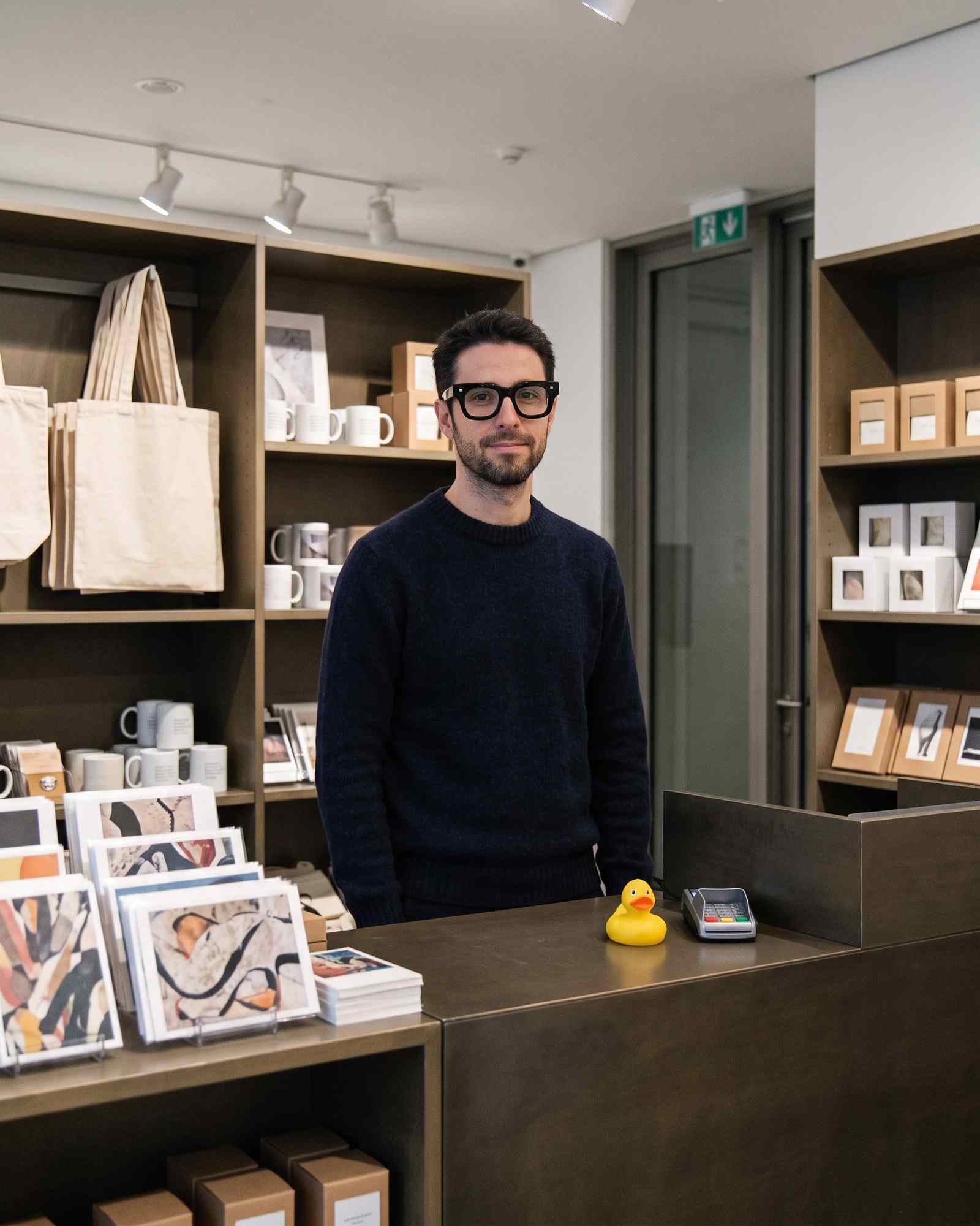 Leo behind the counter of a small museum gift shop with tote bags, mugs, postcards and a rubber duck.